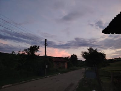 The image depicts a rural scene at dusk in the village of Borintsi, Bulgaria. A paved road runs through the center, leading towards a small brick house with a tiled roof, partially obscured by dark green foliage on either side. Overhead, power lines crisscross the sky, supported by a utility pole. The sky is a gradient of pale blue and purple, with wisps of clouds tinged with pink from the setting sun. Silhouetted against the sky on the right side of the frame is the dark shape of a tiled roof, possibly belonging to a building just out of view. The overall atmosphere is tranquil and subdued, characteristic of twilight in a rural setting.