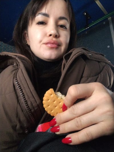 A woman in a brown puffer jacket and black turtleneck holds a cracker with filling. She has a nose ring, dark hair with bangs, and is looking at the camera. Her fingernails are painted red. The background appears to be the interior of a vehicle, possibly a bus.