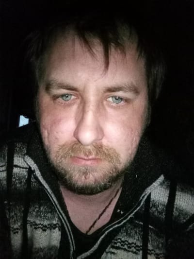 A close-up, dimly lit portrait of a middle-aged man with light blue eyes. He has light brown hair with some receding at the temples, and a beard that is light brown with reddish undertones. His skin appears textured, with some visible scarring. He is wearing a dark-colored shirt under a dark gray and white patterned hooded sweater with a zip-up style. The background is dark and mostly obscured. This does not have any information to confirm the location in the city of Parabel, Russia.