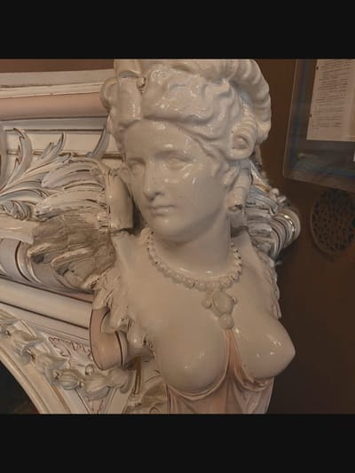 A close-up, slightly tilted shot shows a white porcelain bust of a woman wearing a pearl necklace. The bust is part of a larger decorative piece that includes ornate, carved elements. The woman's hair is styled in curls, and she has a delicate facial expression. The background is a dark, textured surface, with a framed document and a decorative circular object visible to the right. The overall scene appears to be an interior setting, possibly a museum or antique shop. There is no indication of Saint-Août, France in the image.