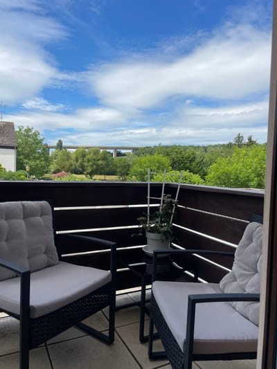 The image shows a balcony overlooking a lush green landscape with a large bridge in the distance. The balcony has two comfortable chairs with light gray cushions and a small side table. A potted plant sits on the table. The sky is blue with scattered white clouds. The scene suggests a peaceful, outdoor setting, likely in Alzey, Germany, given the architecture and vegetation.