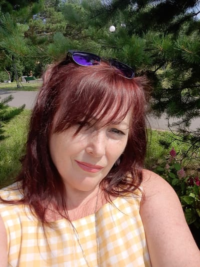 A woman with auburn hair and freckles is taking a selfie in a park. She is wearing a yellow and white checkered dress and a silver necklace. Sunglasses are perched on top of her head. She is smiling and looking at the camera. Behind her, pine trees and green grass are visible. The scene appears to be outdoors in a park setting, possibly in Зеленогорский, Russia, given the context provided.