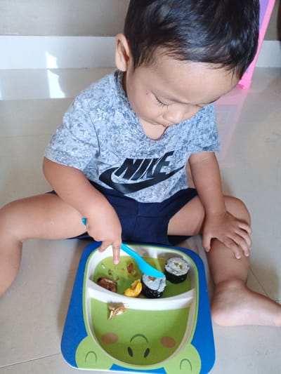 A young child, with dark hair and wearing a gray t-shirt with a black Nike logo and blue shorts, sits on a tiled floor in Cibungbulang, Indonesia, looking down at a compartmentalized plate with a frog design. The plate contains pieces of sushi and other food items. The child is holding a blue fork, about to pick up a piece of sushi. The background shows a white wall with a shadow, and a pink object is partially visible to the right. The lighting suggests an indoor setting with natural light.