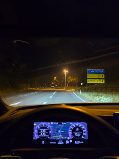 The image shows a first-person view from the driver's seat of a car at night, driving on a road. The headlights of the car illuminate the road ahead, which has white lane markings. There are streetlights visible in the distance, illuminating trees and vegetation on the side of the road. On the right side of the road, there are three German road signs. The top sign is blue with white arrows and numbers "3 67". The middle sign is yellow with "Rüßelsheim" and smaller text "43 519" with a right arrow. The bottom sign is yellow with "Frankfurt 19 km" and "Raunheim 3 km" with right arrows. The dashboard of the car is visible in the foreground, with a digital display showing navigation information and vehicle metrics. The scene is consistent with driving on a road in or near Flörsheim, Germany, based on the road signs indicating directions to Rüßelsheim, Frankfurt, and Raunheim, all of which are in the vicinity of Flörsheim.