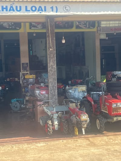The image captures a storefront in Trung Hồ, Vietnam, which appears to be a dealer of agricultural machinery. A large sign above the entrance reads "KHÂU LOẠI 1)" in blue lettering. A telephone icon is also visible on the sign.
Inside the dimly lit shop, various agricultural machines are displayed. In the foreground, two red multi-purpose tractors are prominently featured. The tractor on the right has "YN 20100" written on its front. To the left of these tractors, a turquoise water pump is visible. Further back, other machinery, including red engines and tires, are stored. The environment suggests a commercial setting for machinery sales.