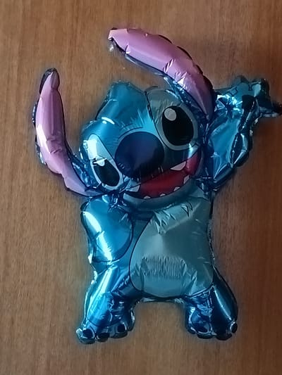 A shiny, blue, foil balloon in the shape of Stitch, a character from Disney's Lilo & Stitch, is attached to a wooden surface. The balloon is inflated and positioned vertically, with its head at the top and feet at the bottom. Stitch's arms are raised in a playful pose, and its mouth is open in a wide smile, revealing sharp teeth. The balloon's surface reflects light, creating highlights and shadows that give it a three-dimensional appearance. The wooden surface appears to be a wall or a door, with a visible grain pattern. There are no people or other objects in the immediate vicinity of the balloon. The scene is indoors, with no visible windows or natural light sources. The location is not specified within Bückeburg, Germany.