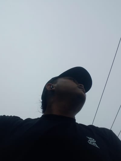 A low-angle, medium shot captures a person from the chest up, looking upwards towards an overcast sky. The individual is wearing a black t-shirt with a light blue logo on the left chest and a black baseball cap. A single black earbud is visible in their right ear. The background consists of a uniformly grey sky and two dark, parallel lines that appear to be power lines or cables. The overall impression is one of a casual outdoor moment, possibly on a cloudy day in a location with overhead wires. The provided location information suggests a possible urban or semi-urban setting within Kubang Jaya, Indonesia, though the image itself does not contain specific identifying markers for this location.