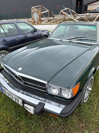 An overhead shot captures the front of a dark green Mercedes-Benz SL convertible parked on a grassy area in Natakhtari, Georgia. The car's hood and windshield are the primary focus, with a portion of a dark blue hatchback visible to the left. In the background, a dark industrial building and rusty metal structures are seen. The license plate on the Mercedes reads "NU-812-VV". A small white object with a checkered flag design is attached to the front bumper. The scene appears to be outdoors, possibly in a parking lot or a yard.