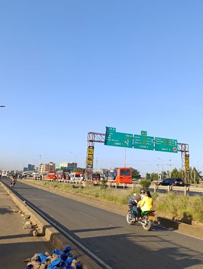 An urban scene shows a multi-lane highway bustling with traffic, including buses, cars, and motorcycles. Overhead signage indicates "EXIT 9" to "tharai" and "Nairobi" as destinations. The signage also provides distances in kilometers to Eldoret and Kisumu. The sky is clear and blue. A motorcycle with two passengers is in the foreground. A pile of blue shoes lies on the side of the road. A sign reading "USE LOW BEAM" is seen above the road. Another sign reading "SPEED KILLS" is visible on the right.