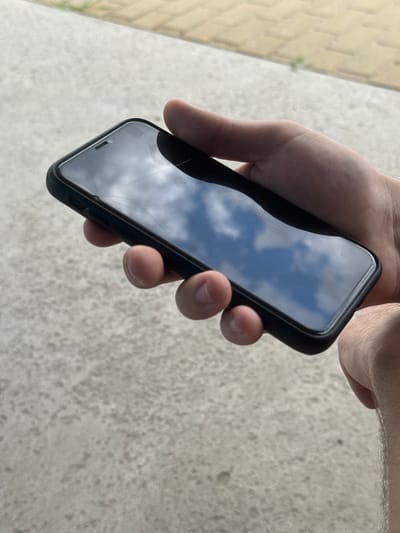 A close-up, overhead shot shows a person's left hand holding a black smartphone. The phone is oriented horizontally with the screen facing upwards, reflecting a blue sky with white clouds. The screen appears to have some minor scratches. The background consists of a textured, light grey surface, possibly concrete, with a hint of a yellow tiled path or wall in the upper right corner. The lighting suggests it is daytime. The location is identified as Shulhynka, Ukraine, though no specific landmarks are visible to confirm this.