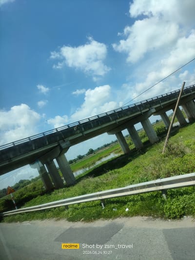 An elevated concrete bridge, supported by numerous pillars, spans across a green, grassy landscape with a body of water visible in the background. The sky above is a bright blue, dotted with scattered white clouds. A metal guardrail runs along the edge of the road in the foreground. A yellow triangular warning sign with a black border and a red vertical stripe is visible on the left side of the image. The scene is captured from a low angle, suggesting the photograph was taken from a moving vehicle. The timestamp "2026.06.24 10:27" is visible in the bottom right corner. The location is specified as Gò Dầu, Vietnam.