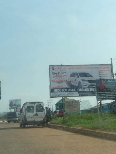 In Haifam, Nigeria, a street scene unfolds under a clear sky. A white van, appearing weathered and possibly used for public transport, is parked on the right side of the road. Its rear doors are slightly open, revealing shadowy figures inside. Beside the van, a person in a purple shirt and black pants stands near another individual dressed in a dark outfit. In the background, a black SUV is visible on the road, and further back, a red vehicle is parked.
Dominating the mid-ground are several billboards. One large billboard advertises "Saferoute Travels & Logistics" and "Travel With Safe Route," featuring a white minivan. Another billboard displays a red handprint and text related to empowering women and children. A smaller billboard for "Goodwill" shows portraits of three men in traditional attire.
The environment is characterized by a paved road with a concrete barrier separating it from a grassy embankment. Beyond the embankment, structures with corrugated metal roofs and various buildings are scattered across the landscape. The overall impression is one of a functional, if somewhat unkempt, urban or semi-urban environment in Nigeria.