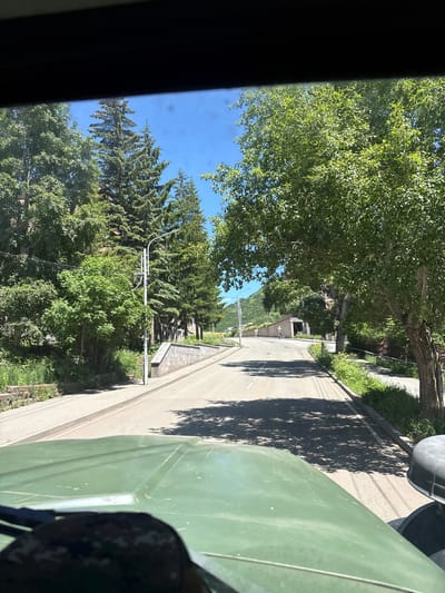 The image depicts a view from inside a vehicle, looking out onto a paved road in Jermuk, Armenia. Tall evergreen trees and deciduous trees line both sides of the road. A modern street lamp stands on the right side of the road. In the distance, the road curves and ascends towards a building with a tunnel-like entrance. The sky is clear and blue, indicating a sunny day. The hood of the vehicle is visible in the foreground.