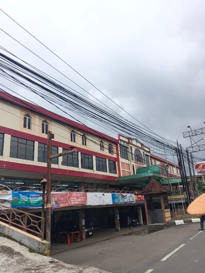 An outdoor urban scene depicts a multi-story commercial building under a cloudy, overcast sky, located in Palayan, Philippines. The building's facade is primarily cream-colored with red horizontal bands, featuring dark-tinted windows on the second floor and smaller, arched-top windows on the upper floors. A central section of the building has a more elaborate design with an arch motif and a green awning extending outwards.
Numerous black electrical or communication cables crisscross the upper portion of the frame, spanning diagonally across the sky and in front of the building.
The ground level features an asphalt road with a white edge line in the foreground. To the left, a concrete ramp with a brown metal railing ascends towards the building. Below the second floor, an open-air undercroft area is visible, containing tables, chairs, and a dark sedan parked. Multiple promotional banners with text and graphics, including one blue banner mentioning "Rp.1.020.000," are strung horizontally across the lower part of the building's facade.
A small, brown, hut-like structure with a pointed roof stands on the right side of the street, near the building. On the far right, a metal pole structure, possibly a streetlight, is visible, partially obscuring a red circular sign.
Two individuals are present: one person in a dark shirt and light shorts walks from left to right across the street in the midground, under the building's overhang. A second person, wearing blue pants and holding a light-colored object, is partially visible on the far right edge of the frame, facing away from the viewer.