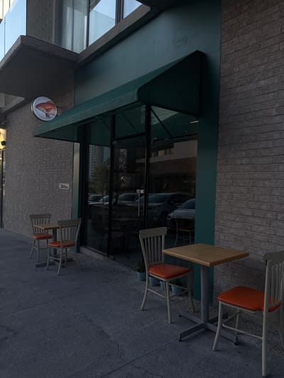 The image shows the exterior of a restaurant in Ciudad Santa Catarina, Mexico. Located at latitude: 25.658, longitude: -100.439, the restaurant has a green awning and a large glass storefront. The storefront reflects images of parked cars. Four tables and chairs are situated on the sidewalk, with a table with two chairs in the foreground and another table with two chairs further back. The table in the foreground has a brown tabletop and orange cushioned chairs. The chairs and tables are grey and have a slatted design. The restaurant's sign is visible, with a depiction of a red fish. The building's exterior is made of brick.