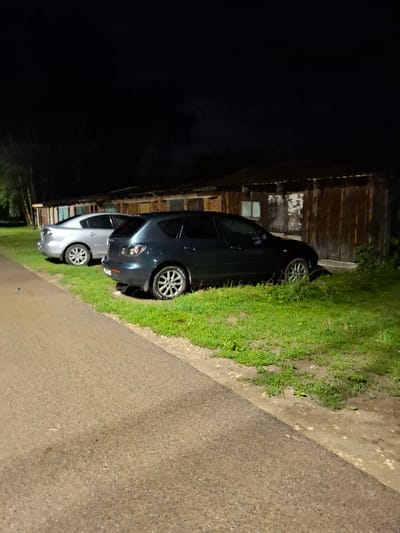 Two parked cars are visible on a grassy area next to a dirt road at night. The car in the foreground is a dark-colored hatchback, and behind it is a silver sedan. The cars are parked in front of a row of rustic wooden structures. The scene appears to be in a rural or semi-rural setting. The image is dimly lit, suggesting it was taken at night, likely with artificial lighting from an unknown source. The location is identified as Novlyanka, Russia.