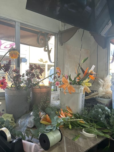 The image depicts a floral shop in Pozos, Costa Rica, located at the coordinates 9.951 latitude, -84.194 longitude. The interior of the shop is a small, enclosed space, likely a converted van or truck, with white walls and a large window. The shop is currently closed for business, with an array of fresh-cut flowers on the counter. Two large grey buckets filled with flower stems rest on the counter. A large white bucket containing a bouquet of orange and yellow flowers is also present, along with a variety of other smaller, loose floral stems. Additional flower stems are strewn on the counter, including green ferns and a small bouquet of white flowers. Green ribbon spools and a roll of tape lie on the counter, suggesting the shop owner is preparing to create floral arrangements. The shop is well-lit, with sunlight entering through the large window.