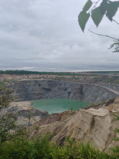 The image depicts a large, open-pit mine with steep, layered walls. A body of turquoise water is visible at the bottom of the pit. The surrounding landscape features sparse vegetation on the slopes, with a dense line of trees visible in the distance. The sky is overcast with gray clouds. The location is identified as the Pioner city, Russia. The scene is a geological or industrial site, possibly a quarry or mine. No people or animals are visible.