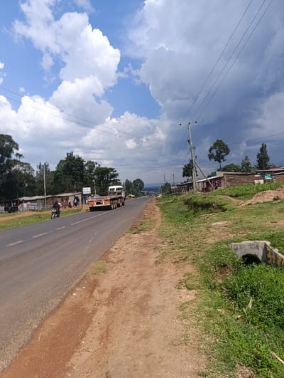 This image depicts a two-lane asphalt road in Kabuchai/Chwele ward, Kenya, under a dynamic sky featuring bright white cumulus clouds on the left and darker, heavier clouds on the right. The road, marked with a faded yellow center line, extends into the distance, gently curving to the right and descending.

On the left side of the road, a narrow dirt shoulder separates the pavement from a series of structures, including single-story buildings with corrugated iron roofs and what appear to be small shops. Beyond these structures, a dense line of trees and bushes is visible. A person on a black motorcycle is traveling towards the viewer on the left lane, while several indistinct figures are observable near the roadside structures further back.

On the right side of the road, a wider, reddish-brown dirt shoulder is present. Beyond this, a grassy bank slopes upwards, featuring a concrete drainage culvert in the immediate foreground. Utility poles with multiple power lines run parallel to the road on this side. Further down the bank, more scattered structures are visible.

A large flatbed truck with multiple axles, light-colored and carrying an unspecified load, is traveling away from the viewer on the right lane of the road. The overall environment suggests a semi-rural or peri-urban setting with a mix of natural vegetation and human habitation.
