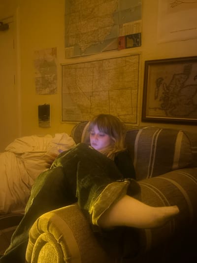 The image captures an indoor scene, likely a living room or bedroom, characterized by warm, dim, yellowish lighting, suggesting evening or nighttime. A young individual with light-colored hair is the central subject, reclining on a striped sofa. Their face is illuminated by the glow of a screen device, indicating they are engrossed in it. The person wears dark trousers or jeans, and one foot, clad in a white sock, is casually propped up on the sofa. A rumpled white blanket or duvet is visible beneath them.
The wall behind the person is decorated with several maps. A large, framed, antique-style map, possibly of Europe, hangs in the upper right. Below it, a large unframed map, partially revealing the text "UNITED STATES," is pinned to the wall. Adjacent to this, a smaller, folded map or booklet is visible, clearly displaying "AUTO MAPA" and "PORTUGAL," with "FORMA PORTUGAL" also decipherable. Another small map is visible lower on the wall, along with a small, dark rectangular object, possibly a speaker or decorative item on a shelf.
The scene depicts a moment of quiet relaxation, with the person focused on their device amidst a collection of geographical wall decor, implying an interest in travel or cartography. The visible text explicitly mentions "PORTUGAL" and "UNITED STATES."