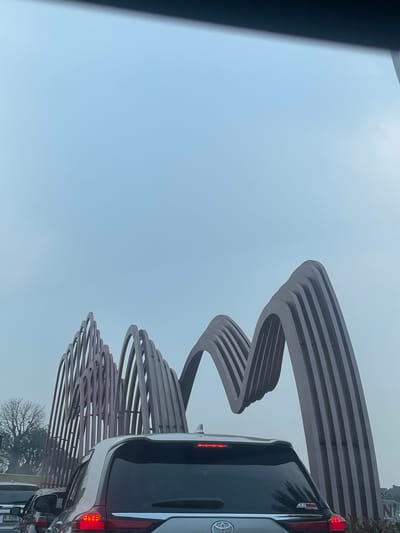 The image depicts a traffic scene in Cijantra, Indonesia, with a distinctive modern sculpture as a backdrop. The sculpture, made of ribbed metal arches, is prominent in the upper half of the frame against a hazy, pale blue sky. In the foreground, several cars are stopped in what appears to be moderate traffic. The rear of a silver SUV is the most visible vehicle, showing its brake lights illuminated. To the left, another vehicle is partially visible with a license plate that reads "A 3706". A bare-branched tree is visible on the left side of the frame, behind the traffic and near the base of the sculpture. The overall atmosphere is overcast, suggesting either early morning, late afternoon, or a day with low cloud cover.