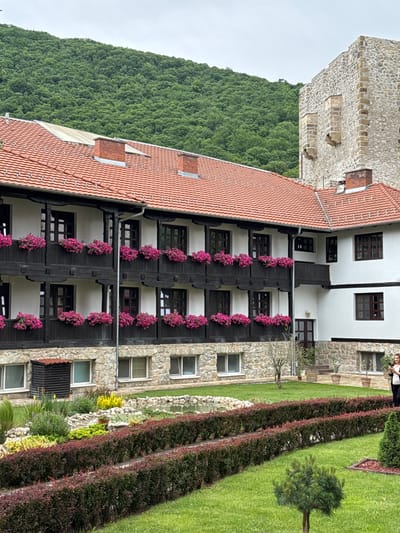 The image shows a building in the city of Despotovac Municipality, Serbia. The building is multi-story and has a combination of stone and white-painted sections with dark wood balconies adorned with pink flower baskets. A stone tower is attached to the building. In the foreground, there is a well-maintained garden with hedges and a small tree. A forested area is visible in the background.