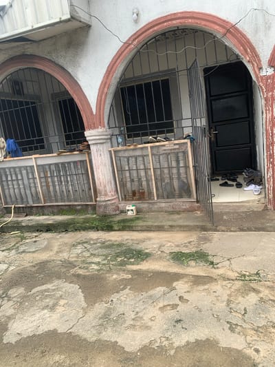 The image shows the exterior of a building with arched doorways and barred windows. The building appears to be made of concrete with reddish-brown trim around the arches. In front of the building, there is a concrete pathway with some moss and cracks. Various items are scattered on the pathway and near the entrance, including shoes, a blue cloth, a green plastic container, and a yellow hose. A black metal door is visible on the right side of the image. The overall impression is of a residential or commercial building in a somewhat neglected state, situated in a Nigerian city.