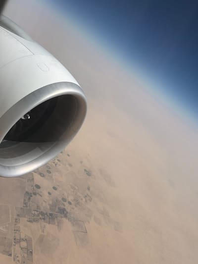 The image shows an aerial view from an airplane. The foreground features the white and silver engine nacelle of the aircraft, partially obscuring the view of the landscape below. The sky transitions from a deep blue at the top left to a hazy, pale orange towards the right and bottom.

Below the sky and aircraft engine, a vast, arid landscape is visible, suggestive of a desert region. The terrain is predominantly sandy and appears sparsely vegetated. Scattered across the landscape are numerous circular and rectangular patterns, indicative of agricultural irrigation systems. These fields, likely under cultivation, stand out against the dry, sandy surroundings. Some of these patterns are distinctly circular, suggesting center-pivot irrigation, while others are rectilinear, indicating different farming methods or land divisions.

The time of day is suggested by the lighting and the gradient of the sky; the light appears to be from a low sun, possibly early morning or late afternoon. The haze in the atmosphere could be due to dust or pollution. The location cues of arid land with organized agricultural plots in such an environment point towards an area where water resources are carefully managed for farming. This is consistent with the provided location context of As Sarrar, Saudi Arabia, an area known for its desert climate and agricultural projects. There is no visible text in the image.