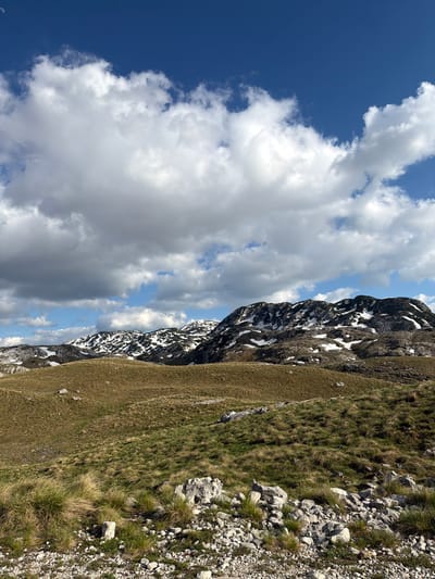 The image shows a mountain landscape in Boričje, Montenegro. The sky is partially covered with clouds, with patches of blue sky visible. The mountains are rocky, with some snow still present. The foreground consists of grassy terrain with rocks and sparse vegetation. There are no people visible in the image.