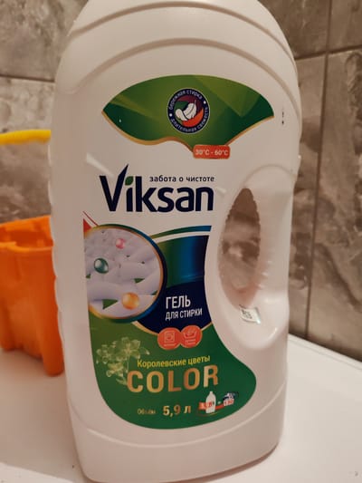 The image shows a white plastic bottle of laundry detergent. The bottle has a green and blue label with text and graphics. The text on the label includes brand names and washing instructions. The bottle is placed on a white surface, possibly a washing machine. There is an orange plastic object to the left of the bottle, and a tiled wall in the background. The image does not contain any information about the city of Zhabinka, Belarus.