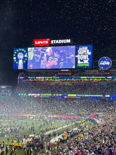 A nighttime scene at Levi's Stadium captures the Seattle Seahawks Super Bowl celebration. The stadium's screen displays an image of the team and the words "Super Bowl Champions." Confetti fills the air as fans pack the stadium. The field is visible with players and personnel. Banners displaying "Seattle Seahawks Super Bowl Champions" are around the stadium. The New England Patriots are also represented on the banners. The stadium's name is prominent.