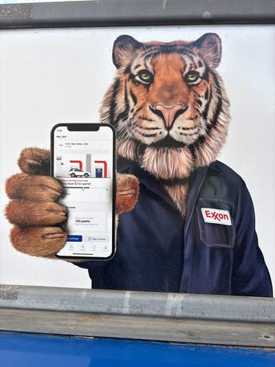 The image shows a large advertisement featuring a tiger wearing a navy blue work uniform with an "Exxon" patch on the chest. The tiger is holding up a smartphone in its paw, displaying a screenshot of what appears to be a mobile app. The app screen shows a gas station location at "1234, Main Street, USA" which is 1.1 miles away. It also displays a message about redeeming rewards for $3 to spend and mentions 100 points. The bottom of the app screen has navigation icons for "Stations," "Rewards," "Offers," and "Account." The advertisement is likely located outdoors, as suggested by the textured white background and what appears to be a metallic bar in the foreground. The city of Bangs, United States of America is mentioned in the prompt but no specific visual cues in the image definitively place it there.