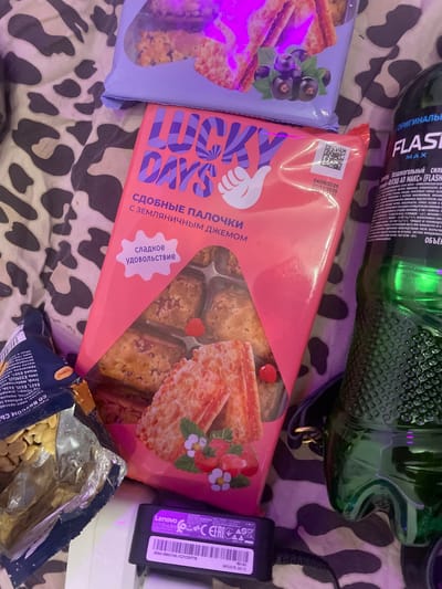 A close-up, top-down view captures a collection of snacks and a drink on a patterned surface, possibly a bedspread. In the foreground, a partially opened bag of peanuts lies to the left, with a pink box of "LUCKY DAYS" cookies, featuring strawberry imagery and Russian text, taking center stage. The cookies are visible through a clear plastic window. To the right, a green bottle of "FLASH MAX" energy drink is prominent, with its label showing Russian text. A black Lenovo power adapter is positioned at the bottom center. The lighting is dim, with a purple hue casting over some of the items. The background exhibits a brown and black camouflage-like pattern. No people or actions are depicted, and the scene is static. The provided image does not contain any information about the city of Zmeinyi, Russia, nor does it offer any clues to suggest this location.