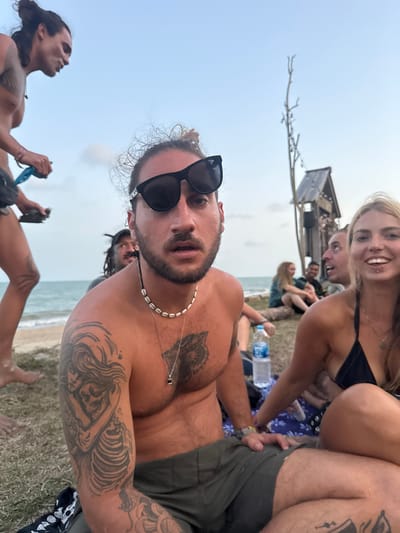 The scene is set on a grassy area adjacent to a sandy beach and the ocean, in Ban Pang Pring, Thailand.
In the foreground, a Caucasian male with dark, curly hair tied in a top knot, a beard, and black sunglasses is positioned centrally, looking directly forward with a neutral expression. He is shirtless, displaying a large wolf tattoo on his left pectoral and an intricate half-sleeve tattoo featuring a skeleton and female figure on his left arm. He wears a white shell necklace and a silver pendant on a thin chain, and dark olive-green shorts, seated on a dark patterned blanket or mat. To his immediate right, a Caucasian female with blonde hair and a lip piercing is partially visible, smiling widely. She wears a black bikini top and a small pendant necklace, also seated on the ground.
In the mid-ground on the far left, a shirtless male with dark hair in a bun stands in profile, looking towards the right, holding small dark objects in his hands. Behind the central male, another male with dreadlocks is partially visible. Further in the background, additional individuals are seated on the ground, appearing to socialize. A clear plastic water bottle is visible on the ground between the central figures and the background group.
The background features the ocean with calm waves under a light blue, hazy sky, suggesting late afternoon or early evening. On the right side of the background, a rustic wooden structure, possibly a hut or small building, stands next to a tall, bare wooden pole or tree sculpture. The environment combines grass, sand, and the sea, indicating a relaxed outdoor gathering.