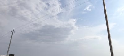 A lone concrete utility pole stands tall in the foreground, angled slightly to the right, against a backdrop of a cloudy sky with patches of blue. Three parallel wires extend horizontally from the top of the pole, disappearing into the distance. In the lower left corner, a smaller, partially obscured sign with Cyrillic text is visible at the base of the pole. To the right, another tall, concrete utility pole, similarly angled, is also visible against the sky. The overall scene suggests an outdoor, possibly rural or semi-urban, environment in Margilan City, Uzbekistan.
