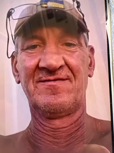 A close-up, head-and-shoulders selfie taken indoors shows a fair-skinned man with short, graying hair, a light beard, and visible wrinkles on his neck. He is wearing a baseball cap with what appears to be a Ukrainian flag emblem. Eyeglasses are perched on top of the cap. The background is a plain, light-colored wall. The image is a photograph of a screen, indicated by the reflections and artifacts on its surface.
