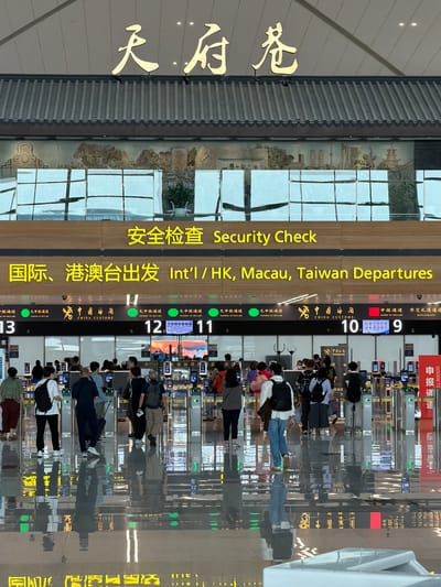 This image captures a bustling airport environment at Chengdu Tianfu International Airport in Jianyang City, China.

At the top of the image, prominent gold Chinese characters spell out "天府空港" (Tianfu Airport) against a plain background, positioned above a large structure featuring a traditional-style dark tiled roof. Below the roof, a decorative mural or relief art piece is visible through a glass partition, depicting a blend of classical Chinese landscapes and modern city skylines with tall buildings and a broadcast tower.

Below this upper architectural element, a horizontal illuminated sign displays "安全检查 Security Check" in yellow text on a dark brown panel. Directly beneath it, another identical sign reads "国际、港澳台出发 Int'l / HK, Macau, Taiwan Departures," also in yellow text on a dark brown background.

Further down, a series of numbered customs or security gates are visible, ranging from 13 on the left to 9 on the right. Above these gates, individual screens indicate different channels: "无申报通道 NOTHING TO DECLARE" with a green light (seen at gates 13, 12, 11), and "申报通道 GOODS TO DECLARE" with a red light (seen at gate 9), and "外交礼遇通道 DIPLOMATS." A large "中国海关 CHINA CUSTOMS" logo is centrally displayed above the gates.

The floor is highly polished and reflective, creating clear inverted reflections of the overhead signs, the people, and the gates. Numerous individuals, predominantly adults, are observed in the lower half of the image. They are engaged in walking towards and standing at the customs gates, appearing to be passengers undergoing pre-departure procedures. Most are carrying backpacks, small bags, or rolling luggage. Their attire is casual to semi-casual. A white or light-colored refuse bin is partially visible in the bottom right corner, also reflecting the ceiling lights.