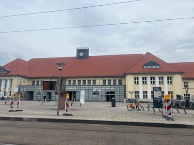 The image shows the exterior of Herne Hauptbahnhof, a train station in Herne, Germany. The building has a red-tiled roof and a prominent clock tower. In front of the station, there is a paved plaza with a tram track in the foreground. Several people are visible walking in the plaza, and there are also electric scooters and bicycles parked. Traffic cones with flashing yellow lights are placed on the plaza, suggesting ongoing construction or maintenance. A digital display board shows train departure information. The sky is overcast.