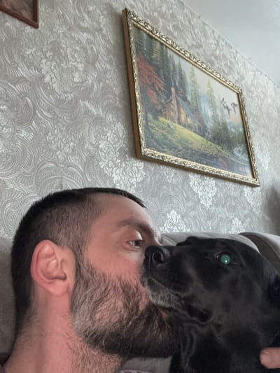 The image portrays an indoor scene in Reftinsky, Russia, featuring an adult male and a black dog. The man, visible from his upper chest to his mid-forehead, has short dark hair, a beard, and brown eyes. His right cheek is pressed against the snout of a black dog, which has short fur and a visible green left eye. The man's gaze is directed slightly upwards and to the right. The background wall is covered in light-colored wallpaper featuring a prominent damask pattern. A rectangular landscape painting, framed in ornate gold, hangs on the wall, depicting a rustic cabin within a forested area, a body of water, and birds in flight. A beige or light brown upholstered surface is partially visible behind the man's head and shoulder.