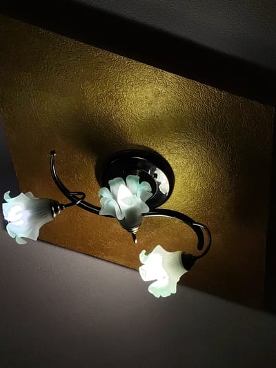 A ceiling light fixture with three frosted glass shades shaped like flowers is mounted on a gold textured ceiling panel. The fixture has a black base and dark metal arms that curve and twist to hold the shades. Each shade emits a soft, white light. The gold panel is angled, with a dark shadow cast across it from above. The wall below the panel is a dark grey color. The image appears to be taken from below, looking up at the fixture and ceiling.