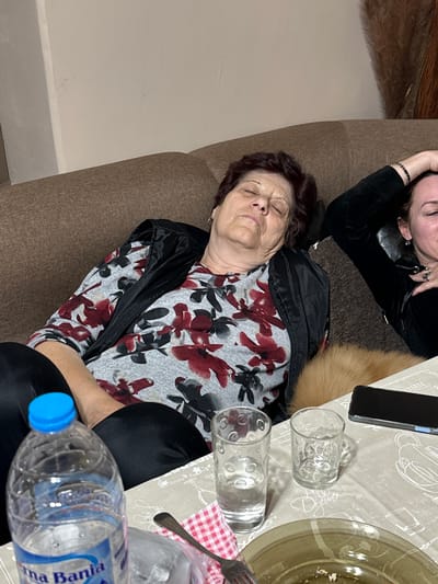 The image shows two women resting on a brown couch adjacent to a dining table. The older woman, with short auburn hair, lies with her eyes closed, wearing a grey floral patterned shirt and a black vest. A second woman with dark hair is partially visible at the top of the frame, also resting. A dining table covered with a patterned tablecloth is visible with a smart phone, water glasses, a plate with remnants of food, a fork on a red and white checkered napkin, and a plastic water bottle. Based on the brand name "Vina Banja" displayed on the water bottle, it is possible the location could be in the city of Vinarovo, Bulgaria.