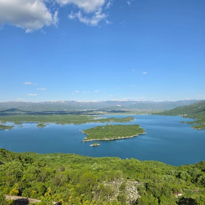 The image shows a high-angle view of the Slansko Lake in Ponikvica, Montenegro, on a clear day. The lake is blue and contains several small, green, tree-covered islands. Lush green forests cover the terrain on the right side of the image. In the distance, mountains are visible and there is a town or settlement located in front of the mountains. The sky is clear blue with a few scattered clouds.
