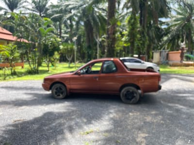 The image captures a side profile of a rusty orange, two-door pickup truck parked on a gravel driveway. The truck has black rims on its wheels. Behind the truck, a white sedan is partially visible, also parked on the gravel. The environment is characterized by lush green vegetation, including palm trees and other foliage, suggesting a tropical setting. In the background, there are several small structures, including a building with a red tiled roof and other weathered-looking buildings. The scene appears to be an outdoor area, possibly a residential or resort property, located in Khao Khram, Thailand, given the characteristic palm trees and tropical vegetation. The overall lighting suggests it is daytime, likely with bright sunlight causing dappled shadows on the ground. There are no people visible in the image.