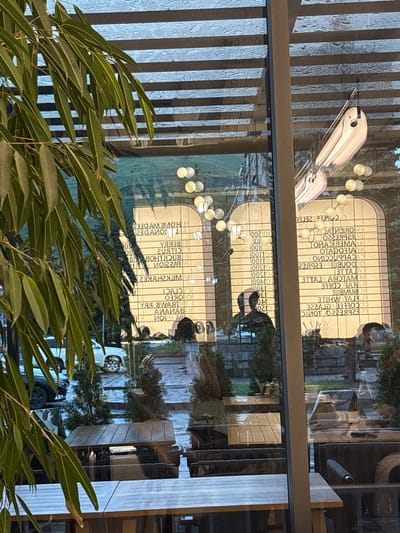 A glass-enclosed cafe's exterior view shows a reflective window with a menu and reflections of trees, cars, and people. The menu, written in Cyrillic and Latin alphabets, displays food and drink items with prices. In the foreground, a branch with long green leaves obscures part of the view. Outside, tables and chairs are visible, some covered by wooden canopies, suggesting an outdoor seating area. A white SUV and a black car are parked on a wet road, indicating recent rain. The lighting suggests it is either dusk or an overcast day. The scene is indoors looking outwards, with the cafe's interior lighting reflecting on the window. The city of Hrazdan, Armenia, is inferred from the context of previous interactions.