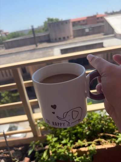 A hand holds a white mug with a cartoon elephant and the text "BE HAPPY" written on it, filled with a light brown beverage, possibly coffee. The mug is positioned in front of a balcony railing, and in the background, there are buildings and a clear blue sky, suggesting an outdoor urban setting. The mug has a brown handle and a white extension where it connects to the mug body. The perspective suggests the photo was taken from a higher vantage point, looking outwards from a balcony.
