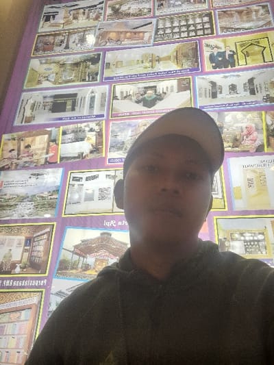 A male individual is centered in the foreground, facing the camera and partially obscuring a wall display. He wears a dark hooded sweatshirt and a light-colored baseball cap reversed on his head. The background consists of a large wall covered with numerous rectangular posters, each framed in yellow and set against a purple background. These posters depict various architectural designs and interior views, including what appear to be religious structures, prayer halls, and library interiors. Some images contain indistinct human figures. Visible text on the posters is primarily in an Indonesian script. The scene is located in the city of Glantengan, Indonesia, and presents as a public information or exhibition display.
