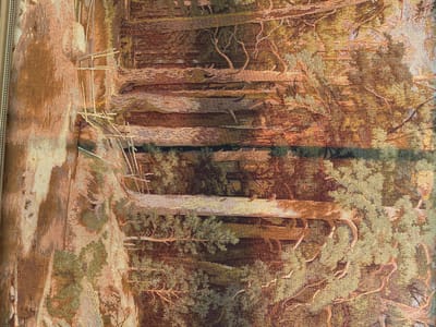 This is a close-up, vertical shot of a tapestry depicting a forest scene. The tapestry is highly detailed, showcasing the textures of tree bark, foliage, and branches. The dominant colors are earthy tones of brown, green, and gold, creating a warm and naturalistic effect. In the foreground, several large tree trunks are prominent, with intricate root systems visible. The background suggests a dense forest with dappled light filtering through the canopy. There appears to be a reflective surface, possibly a body of water, mirroring the trees and creating a sense of depth. A wooden structure, perhaps a bridge or fence, is also depicted within the forest. The overall impression is one of a serene and slightly mysterious woodland environment. The tapestry is framed by a decorative border, visible on the right side of the image. No text or identifiable people are present in the scene. The lighting suggests it could be daytime, with soft, natural light illuminating the forest.
