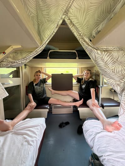 Two young women are in a train compartment. One woman is lying on the lower berth, wearing a black t-shirt and shorts. The other woman is lying on the opposite lower berth, also wearing a black t-shirt and shorts. The compartment has leaf-patterned curtains, and the berths have white sheets. Shoes are visible on the floor. A window with greenery outside is seen. The location is a train compartment, but there's no way to pinpoint the exact location, Eremeevka, Russia, from this image alone.
