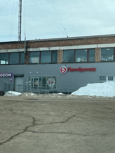 The image displays the exterior of a two-story commercial building, located in Уральский, Russia. The upper floor is constructed of reddish-brown brick, featuring multiple rectangular windows that reveal indistinct interior contents. A corrugated metal roof covers the structure. The lower floor is clad in grey paneling.

The dominant business on the lower level is a "Пятёрочка" (Pyaterochka) supermarket, identified by its red Cyrillic logo with a white '5' in a green stylized leaf. To the left, an "OZON" pickup point is marked by blue and white signage. Next to the Ozon signage is an entrance with a gray metal ramp and stairs leading to a door. Above this entrance, a sign reads "ПАРИКМАХЕРСКАЯ" (Hairdressing Salon) with "2 ЭТАЖ" (2nd Floor). The central entrance features glass double doors, displaying internal signs including operating hours "8-22" and a circular red promotional sign with "30".

The foreground consists of a cracked, rough, grey ground surface, consistent with a parking area or access road. Large piles of white snow are accumulated against the base of the building, notably on the right side. The sky above is overcast and light grey.

A tall, lattice-structured antenna or communication tower is visible behind and above the left side of the building, extending upwards. Several dark birds are perched on the roofline and on a vertical pole attached to the building. No human individuals are present within the frame.