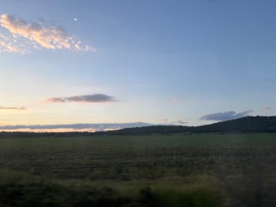 The image displays a rural landscape captured at dusk in Smilets, Bulgaria, observed from a moving vehicle. The upper two-thirds of the frame consist of a sky gradient, transitioning from pale blue in the upper right to soft orange-pink and light blue hues near the horizon on the left. A small, bright moon is visible in the upper central-left sky. Scattered clouds are present; a prominent, elongated formation of light-colored, wispy clouds, illuminated with golden-pink tones, spans the upper left quadrant. Smaller, darker clouds are visible towards the right and along the lower horizon line. Rectangular reflections or window distortions are subtly visible in the upper right quadrant of the sky. The middle ground is dominated by a dark green, undulating line of densely forested hills or mountains that form the horizon. Below the treeline, an extensive area of green cultivated fields stretches across the landscape. Several indistinct dark masses, consistent with hay bales, are faintly discernible in the distant fields. The immediate foreground is blurred horizontally due to motion, showing streaks of dark green and brown vegetation. No people or animals are present in the scene.